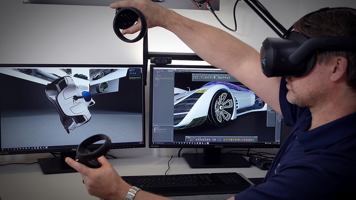 Person wearing a VR headset and holding controllers, in front of two monitors displaying a car design